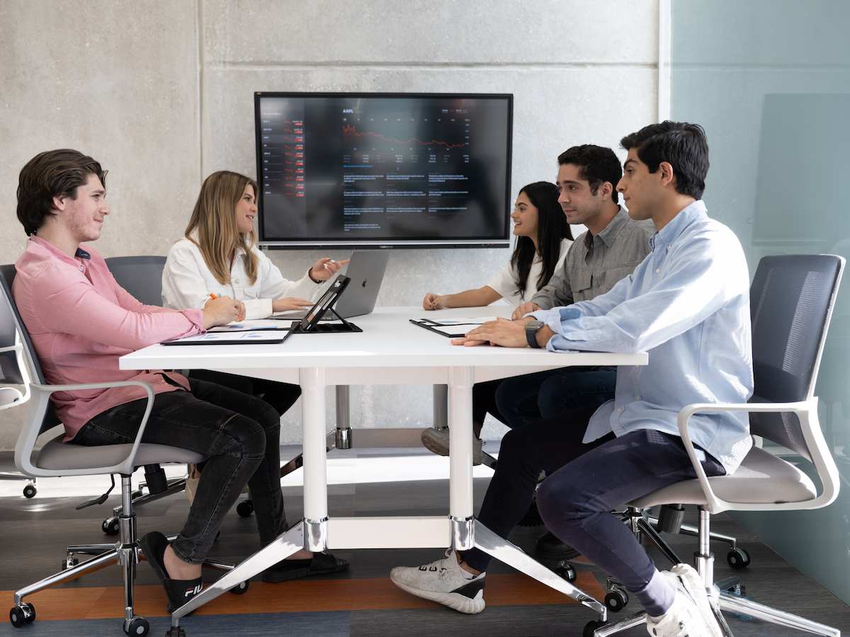 Grupo de cinco estudiantes reunidos alrededor de una mesa, conversando mientras observan datos proyectados en una pantalla de fondo.